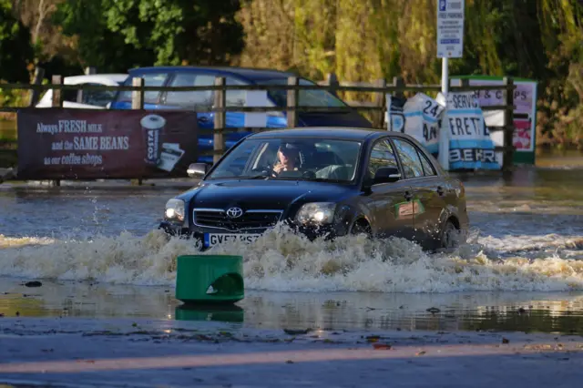 A black car driving through heavily flood waters in Billing Aquadrome. The top of a green bin lid is floating in the water.