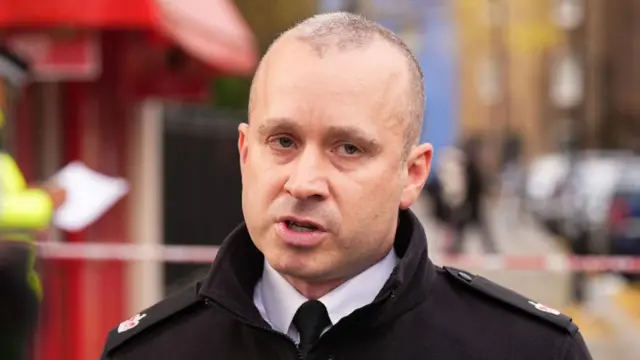 Renowden, a police officer in a black jacket, speaks standing in front of a police cordon across a road