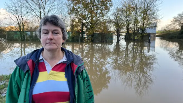 Harriet Balshaw, who is wearing a stripy top and a green jacket, standing in front of flood water