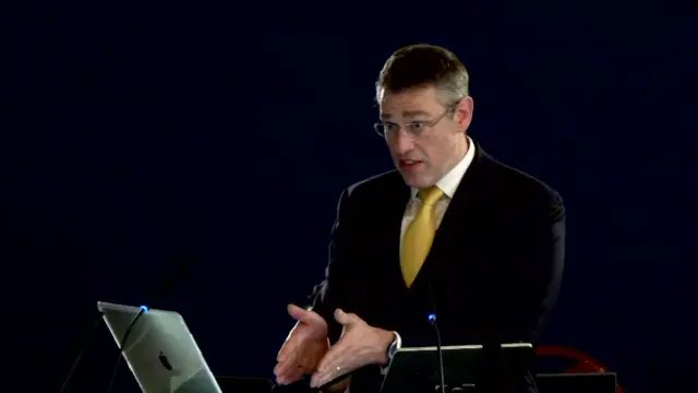 Nick De la Poer stands in a dark suit and yellow tie, wearing glasses and gesticulating with his hands