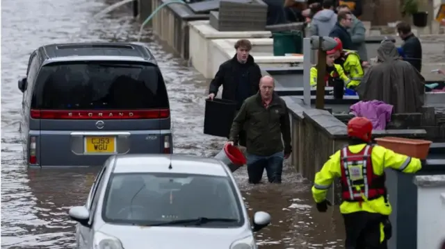 Two men carrying buckets wade through floodwaters near two submerged cars on the road.