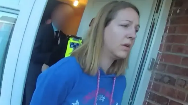 Lucy Letby is led from her house after being arrested by police. She has her hair down and is wearing a blue hoodie. A police officer is behind her.