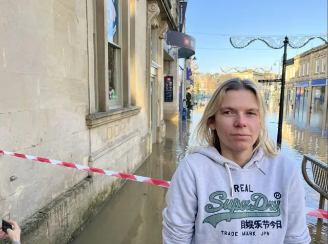 Becky Lyons in a grey Superdry hoodie, stood in front of a red and white warning tape, blocking off a flooded road in Chippenham