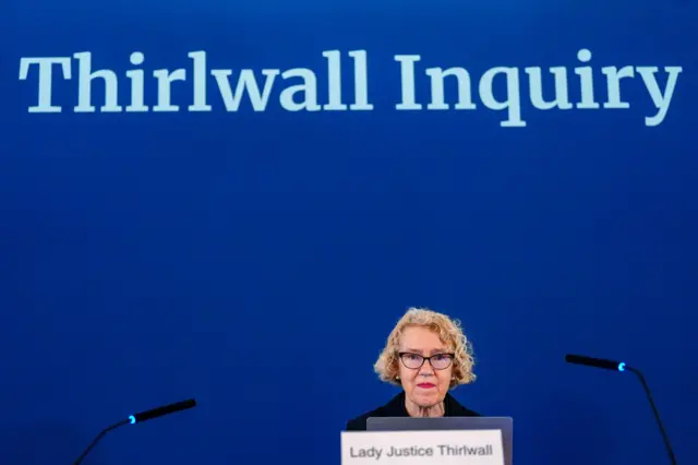 Lady Justice Thirlwall sits behind a white sign with her name on it. She sits in front of a blue background which reads 'Thirlwall Inquiry' in white. She has short, curly blond hair, glasses and is wearing black.