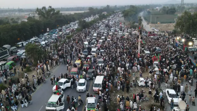 A large crowd, some on foot and some in cars, moving on a highway