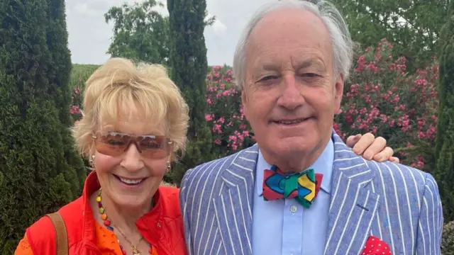 Christine Hamilton is pictured on the left wearing a red jacket and sunglasses. She has her arm around her husband, Neil Hamilton, who wears a stripy blue suit and colourful bow tie