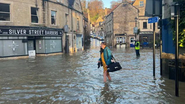 A woman holds up the hem of her yellow dress and holds her shoes as she walks through the flood water.