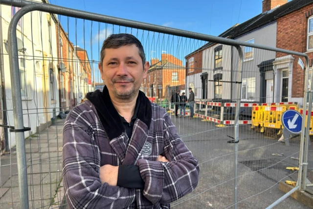 Robert Demeter stood behind a metal fence which has sectioned off a damaged house. He has his arms folded over is chest and looks to be wearing a purple checkered dressing gown.