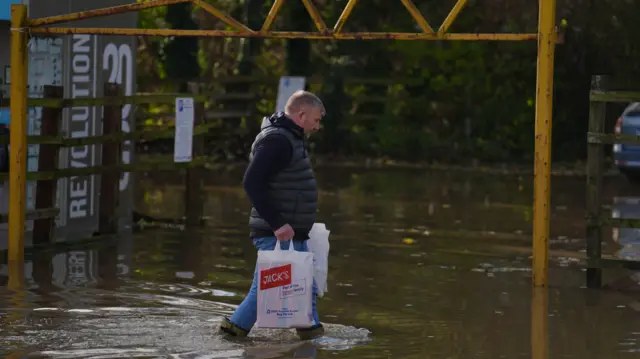 Man walks with shopping bags through flood waters. He is wearing wellies, which the water nearly goes over, and a black gilet.