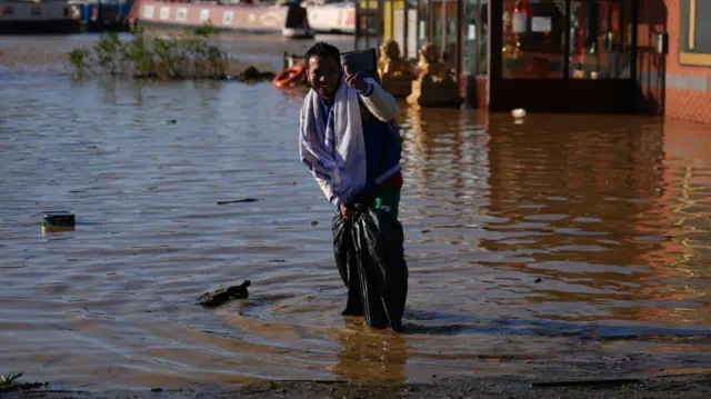 Man gives a thumbs up to the camera while stood in flood water. His clothes are wet.