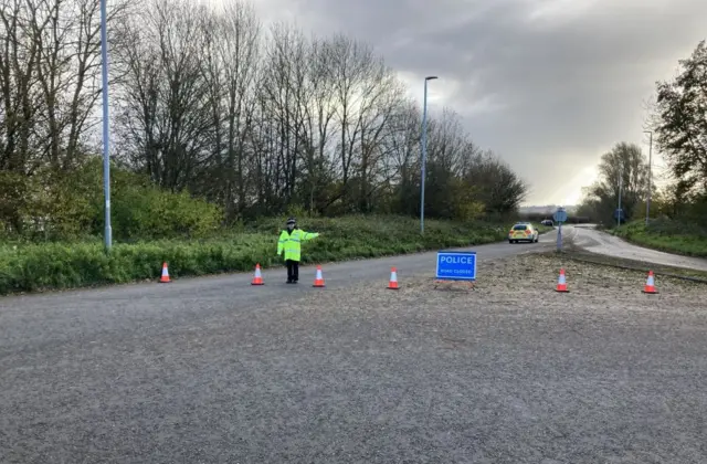 A police officer has cordoned off a road in Ilchester, Somerset, with orange traffic cones and a 'Police road closed' blue sign