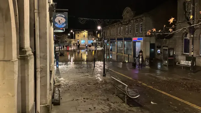 High street scene with shops lining both sides. In the distance the lower part of the high street is flooded from door-to-door. Further up, the road is lined with metal benches and is now flooded.