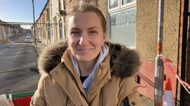 A woman wearing a huge coat smiling at the camera while standing on a residential street