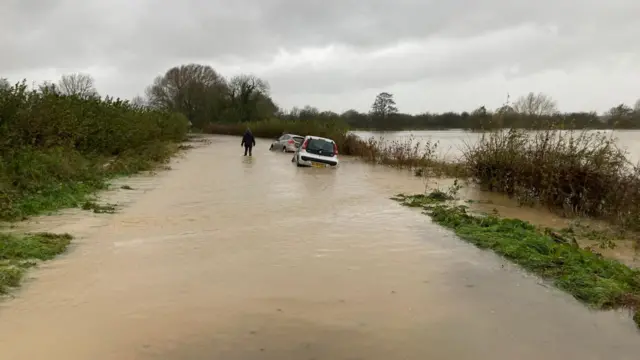 A road is under water and abandoned cars sit to the side. There is a person wading through the water in the middle of the road.