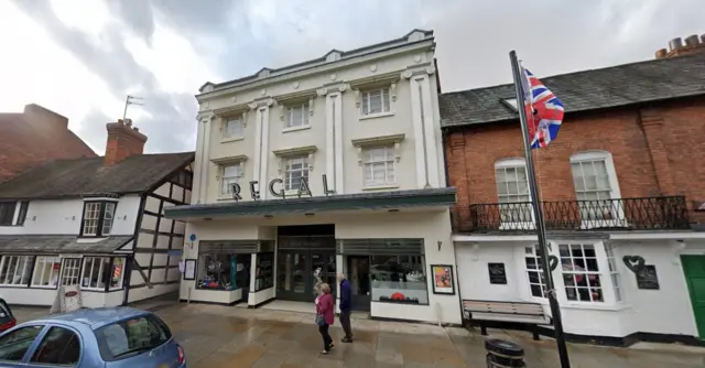 The Regal Theatre is a crea-coloured, three-storey building with a brick-built building to one side and a black and white building to the other side. There is a Union Jack flag on a paved area in front of the building and parked cars.