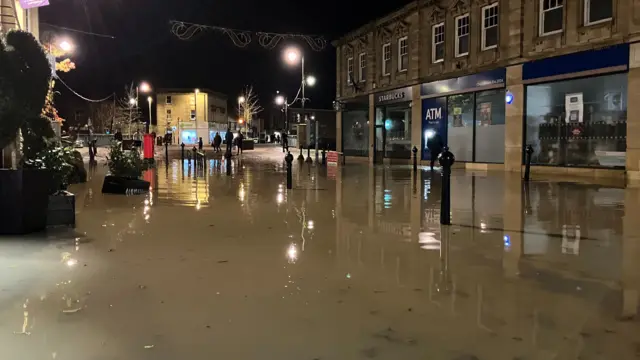 Grey / brown floodwater with black bollards sticking out of it, in an empty shop-lined town centre.