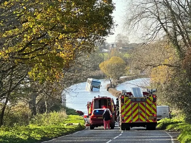 Fire crews in the foreground can be seen attending the scene of a flood where there is a bus stranded in the middle of the water. The fire service vehicles and crews are parked on a section of road that is not underwater.