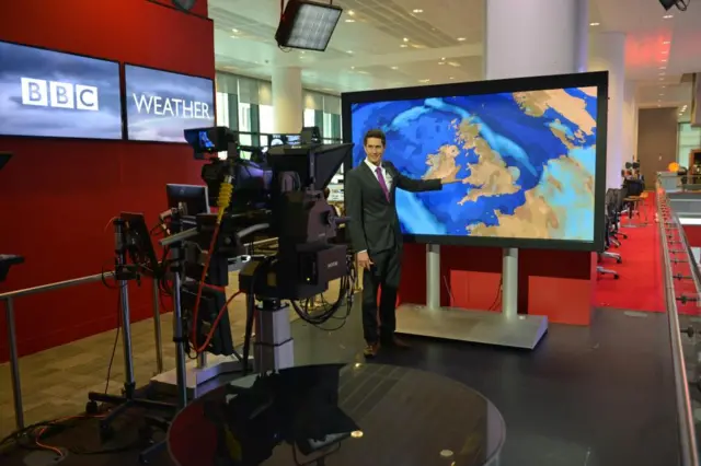 Christ wearing a suit standing in front of a weather map with a camera in front of him