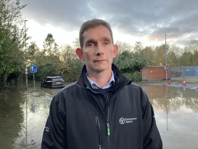 A man in a black Environment Agency jacket stands in front of a flooded car park