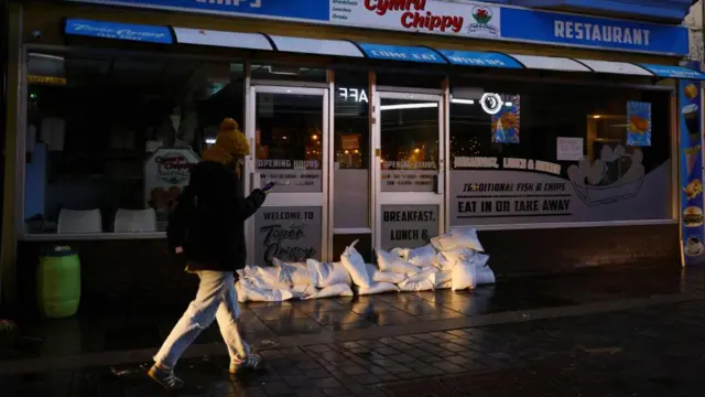 A person walks by a shop with sandbags out front.