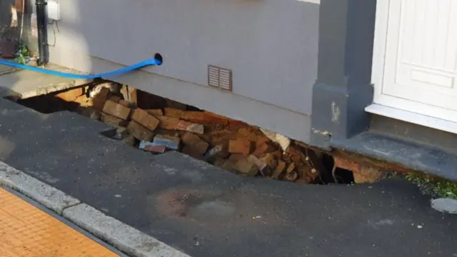 Close up of possible sinkhole outside a house. Loose bricks can be seen below a gap at the bottom of the house. Part of the pavement is no longer complete