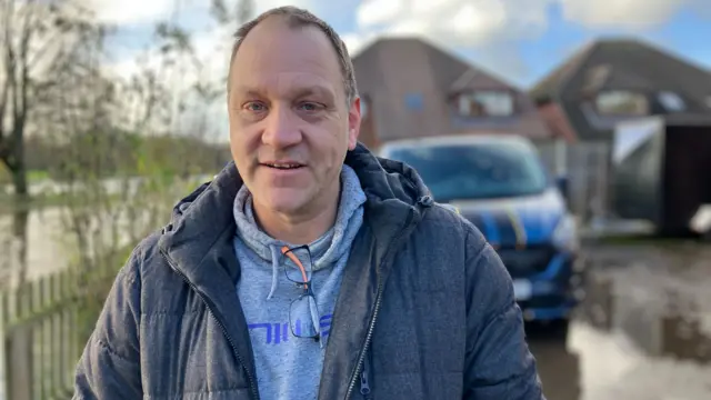 Tall man in grey hoodie with darker grey coat over the top. His is standing in floodwater near his blue van.