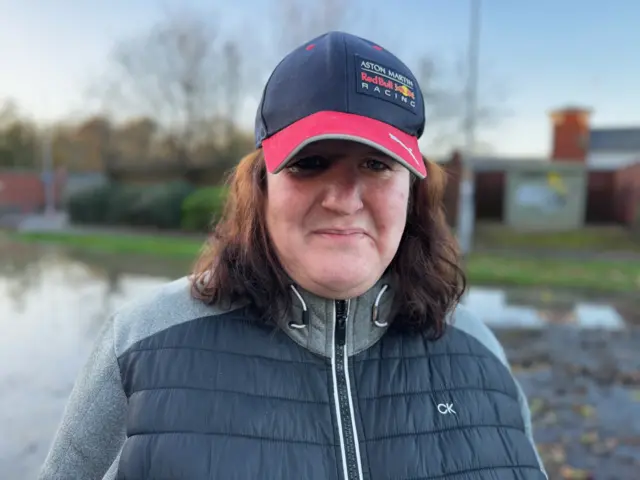 A woman with dark brown hair wearing a blue puffer jacket and red cap looks at the camera.