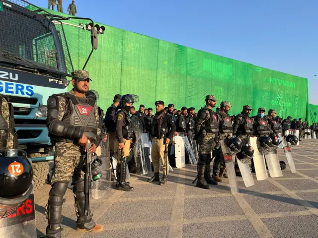 Soldiers with riot shields standing in front of a wall built of containers and some green tarp