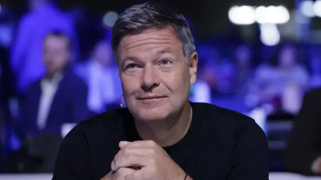 Robert Habeck sits with his hands clasped together looking beyond the camera while smiling. He is wearing a black top and has a microphone attached round his ear. People are sat in the background but are blurred.