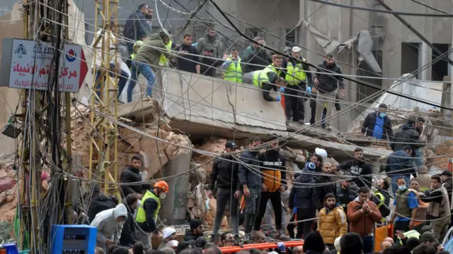 People crowd around a collapsed building. Some men stand atop a fallen concrete pillar