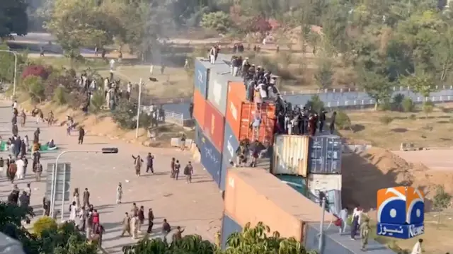 A number of people on top of shipping containers and some on the street