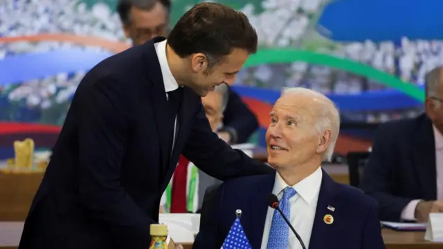 Macron stands beside a seated Biden, his hand on Biden's shoulder, looking down at him as he speaks