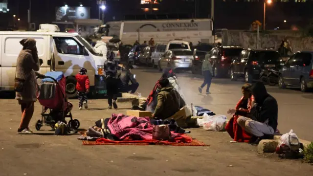 People wrapped in blankets and jackets sit on a footpath. A baby sleeps in a blanket on the floor while a woman stands by a pram