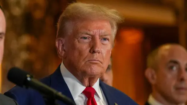 A close up shot of Trump's face, not smiling, as he stands behind a microphone at a news conference at Trump Tower