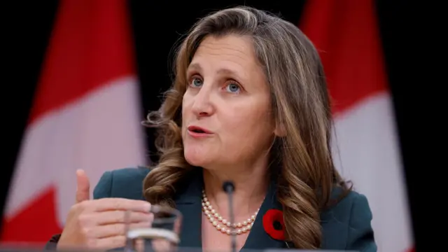 Chrystia Freeland in a blue blazer, sat down and given a statement. There are blurred Canadian flags in the background