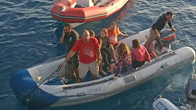 In a picture taken from above, several people are in a dingy, a man in a red t-shirt is stood up at the front holding a rope.