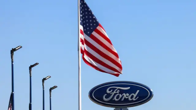 A sign for Ford motor company is at the bottom of the picture against a blue sky. There is an American flag on a flagpole next to it