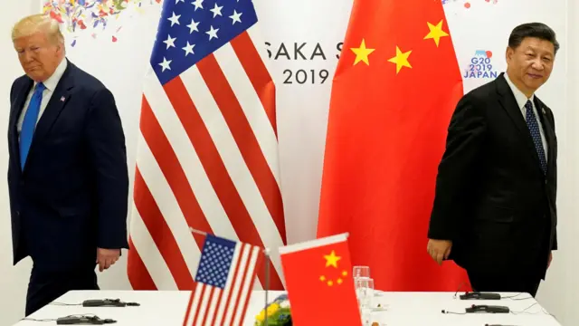 Donald Trump is on the left side of a table in front of an American flag in 2019. On the right is Chinese President Xi Jinping, in front of a Chinese flag