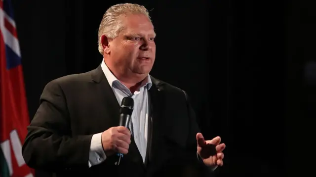 Doug Ford stands on a stage speaking into a microphone, which he is holding in his right hand. He is wearing a suit with a black blazer. He has white hair.