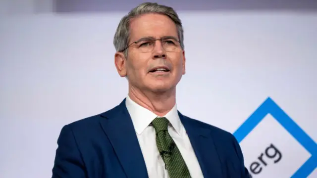 Scott Bessent sat on a stage. He is wearing a suit consisting of a navy blue blazer, white shirt, and dark green tie. He is wearing glasses and has short greying hair.