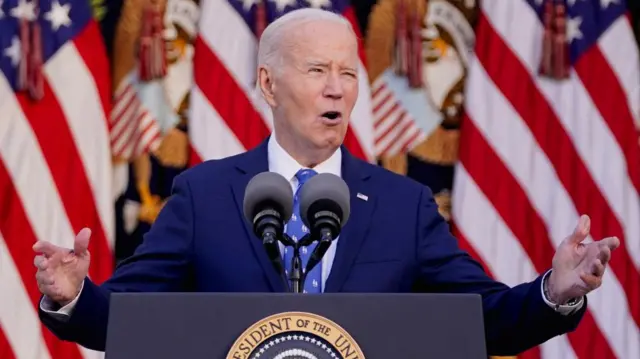 Biden holds his arms out in gesture as he speaks at a presidential lectern in front of US flags