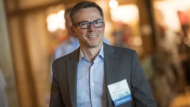 Randall Kroszner walks along while smiling. He is wearing a grey blazer and light blue shit, and has black-rimmed glasses on. He has a badge pinned to his chest that says University of Chicago.