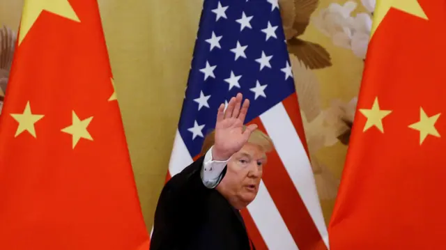 Donald Trump waves his hand at the camera while walking away in front of two China flags and one America flag