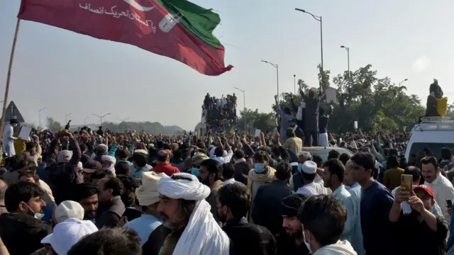 Large crowd standing in the middle of a road, with some cars in the middle