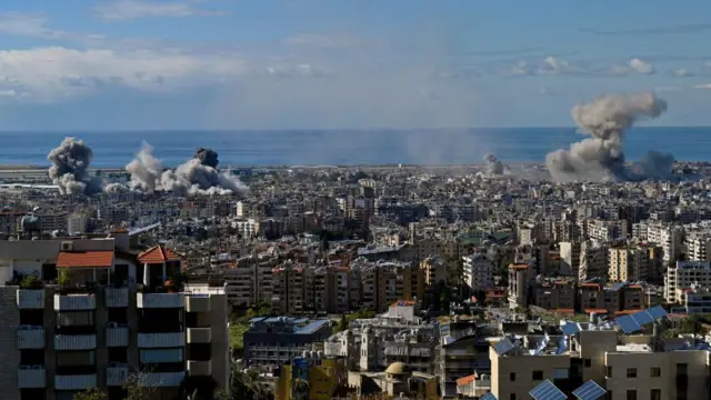 Smoke rises over building that appear to have been hit by Israeli strikes in the southern parts of Beirut. The sky is bright blue and you can see the sand off in the city skyline distance.