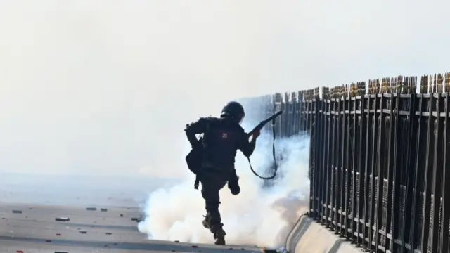 An armed officer kicking what looks to be a can of tear gas