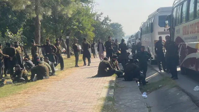 Paramilitary officers standing and sitting on a pavement, next to a line-up of coaches