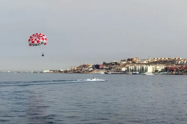 A red and white chcked parachute sails over the sea, there are buildings in the background.-