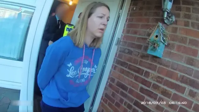 Lucy Letby walking out of house being escorted by police. She is wearing a blue hoodie and has her hands behind her back. One police officer in uniform and a person in a dark suit can be seen walking behind her