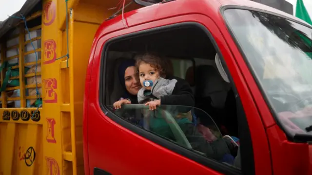 A woman holds a young child, as both look out the window of a large vehicle